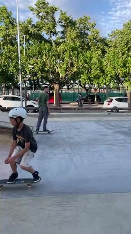 A young boy, wearing a white helmet, dark t-shirt, and white shorts, attempts a skateboarding trick at a concrete skate park. He approaches a low, rusty orange metal rail on his black skateboard with white wheels. As he tries to slide or grind the rail, he loses balance, steps off the skateboard, and lands on his feet next to the rail. The skateboard continues briefly before stopping. He then walks away from the rail and his skateboard. The skate park features various concrete ramps and flat areas. In the background, numerous lush green trees are visible under a bright blue sky with scattered white clouds, suggesting a sunny late afternoon or early evening. Beyond the trees, cars and some buildings can be seen, partially obscured by a chain-link fence.