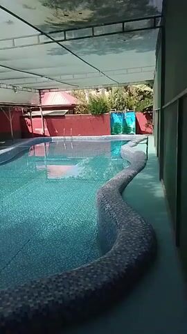 The video pans across an outdoor swimming pool area. The pool is rectangular with a curved edge that is tiled with mosaic patterns. Sunlight streams in from the open side of the covered area. The water is clear and blue, with some ripples on the surface. There are lounge chairs visible under a covered patio area to the left of the pool. To the right, there is a wall with a green and blue graphic. Trees and a red building are visible in the background.