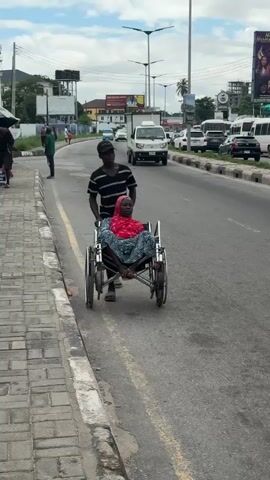This media file shows a man pushing a woman in a wheelchair down a road in Ikoyi, Nigeria. The scene is a busy street with traffic and pedestrians. The man is wearing a striped t-shirt and a black cap, while the woman is wearing a red headscarf and a patterned dress. They are moving from left to right in the frame.

The time of day appears to be daytime, with a cloudy sky suggesting possibly overcast weather. The setting is an urban environment with buildings and billboards visible in the background. One billboard features an advertisement with a person's image. The road has multiple lanes and a yellow dividing line. The sidewalk is made of interlocking bricks.

The activity depicted is the man assisting the woman by pushing her wheelchair. The expressions on their faces are not clearly discernible due to the distance and resolution of the image, but the overall impression is one of movement and perhaps a journey. The presence of other vehicles and people on the street indicates a normal day in a populated area. The image captures a moment of everyday life, highlighting transportation and mobility challenges. There is no discernible text that is clear enough to be read.