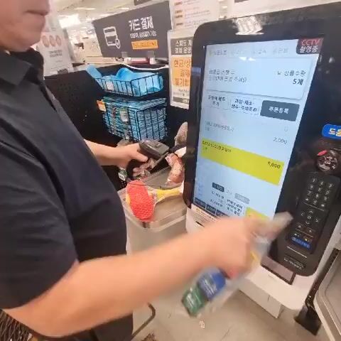 A person in a black short-sleeved shirt and dark shorts is standing at a self-checkout register in a store. The person is holding a barcode scanner in their left hand and a red and yellow mesh bag with an object inside in their right hand. The person is scanning items at the self-checkout machine, which has a large touchscreen display showing a list of items and prices. The background shows shelves stocked with various products. The person appears to be in the process of purchasing groceries or other items.