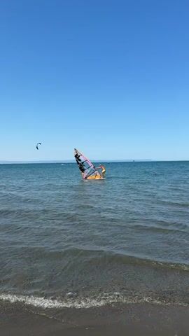 The video shows a windsurfer in the ocean on a sunny day with a clear blue sky. The windsurfer is on a board with a sail. There are two other figures in the distance, one on a kiteboard and another on a paddleboard. The camera is positioned on the shore, looking out at the water. The waves are small and gentle.
