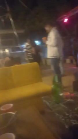 The video shows an outdoor lounge or bar area at night. There are people milling around, and a DJ is playing music at a setup with a screen. The camera focuses on a low table with beer bottles and cups. There are yellow and dark-colored sofas and chairs in the area. The lighting is dim, with blue and white lights illuminating the scene. Some people are standing around talking, while others are walking through the area.