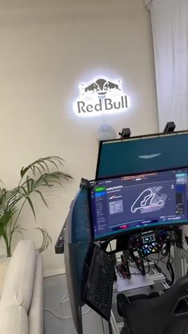 A simulation setup with multiple monitors and a racing wheel is shown. A Red Bull logo is illuminated on the wall behind the monitors. One monitor displays a racetrack layout. Another monitor shows the Aston Martin wings logo. The setup includes a keyboard and pedals. The scene is indoors, possibly a room with a plant.