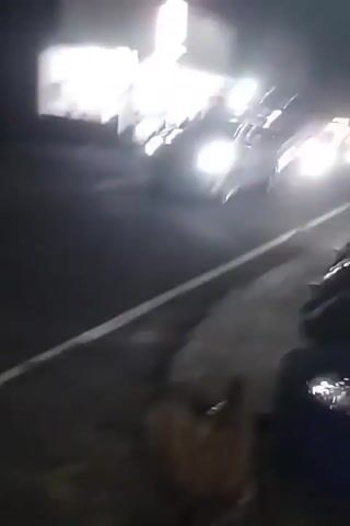 A shaky, nighttime video captures a street scene with several vehicles. In the foreground, a silver car is driving on the road. Further down the road, a motorcycle with two riders passes by, its headlight illuminated. On the left side of the street, there are buildings with lights on, suggesting they might be shops or businesses. A tall lamp post with a bright light is visible in the distance. The road appears to be paved with white lines marking the lanes. The overall atmosphere is dark, with artificial lighting from the vehicles and buildings illuminating the scene. There are no clear indications of any particular event or activity occurring, other than the movement of vehicles on the street.