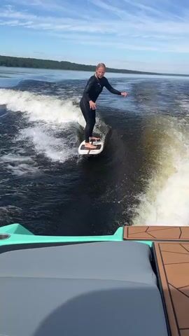 A blonde woman in a black wetsuit surfs a wave behind a boat. The woman is on a white surfboard with black accents. She is wearing black booties. The boat is gray and teal. The water is dark blue with white foam from the wake. The sky is blue with white clouds. There are trees on the horizon. The woman is looking at the camera and smiling. She appears to be standing in a surfing stance with her knees bent. She is positioned on the crest of the wave. The boat's wake is visible to the right of the surfer. The camera is positioned on the boat, looking back at the surfer.