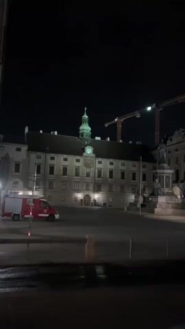 A fire truck is parked in front of a large, ornate building at night. Two construction cranes are visible in the background. The building appears to be a historical landmark, possibly a palace or government building, illuminated by lights. The area in front of the building is a large, open square. A few people are walking in the distance, and a taxi is visible driving away. The ground appears wet, suggesting recent rain.