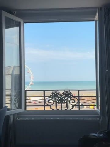 A slightly out-of-focus video shows a view through an open window to the sea and a Ferris wheel. The window is open on the left side, revealing a white window frame and part of a white curtain on the right. A black metal railing with an ornate design is visible across the lower portion of the window. Beyond the railing, a beach and the ocean stretch to the horizon under a clear blue sky with a few wispy clouds. A large white Ferris wheel with red gondolas is visible on the left side of the frame, situated near the beach. The lighting suggests it is daytime. The scene is still and there are no discernable people or actions occurring within the immediate view.
