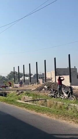 The video shows a construction site with several cinder block walls being built.  There are at least six tall black pillars erected in a line, some of which appear to be part of the structure.  Several individuals are present on the site, engaged in various construction tasks. One person, wearing a red shirt, is operating a wheelbarrow. Another person, in a blue shirt, is also working near the foundation.  In the background, there are houses and trees visible. The ground is a mix of dirt and some grassy areas. A paved road runs along the foreground, with a grassy verge and a drainage ditch between the road and the construction site.  The sky is clear and blue.