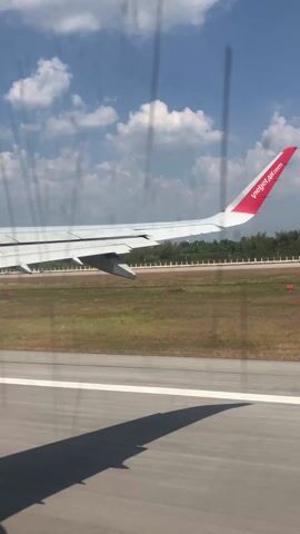 The view from inside an airplane shows the plane landing on a runway. The plane is Vietjet Air and the tail wing with their logo is visible. Through the window, the runway and nearby vegetation are visible, along with a partly cloudy sky.