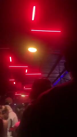 The video opens with a view of red neon lights and a bright overhead light, possibly in a club or entertainment venue. The camera pans down to show the silhouettes of people in the foreground, with other figures and glowing lights visible in the background. The scene is dark and dimly lit, with the red and blue lights creating a vibrant atmosphere. The video then transitions to complete darkness.