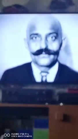 The video shows a television screen displaying a black and white photograph of a bald man with a prominent mustache, wearing a suit and tie. The camera then cuts to a color image of a woman with short, dark hair and sunglasses, wearing a red jacket and a scarf. The background on the television screen is blurry and appears to be a television set with some electronic equipment below it. There is also a watermark at the bottom of the screen that reads "HUAWEI P30 PRO LEICA QUAD CAMERA".