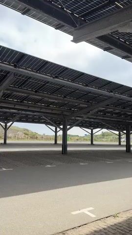 The video shows a parking lot covered by solar panels. The camera pans across the parking lot, showing rows of parked cars under the solar panel structures. The solar panels are mounted on metal frames supported by columns. The ground of the parking lot appears to be a combination of asphalt and permeable paving blocks. In the background, there are some sand dunes and vegetation. The sky is cloudy.