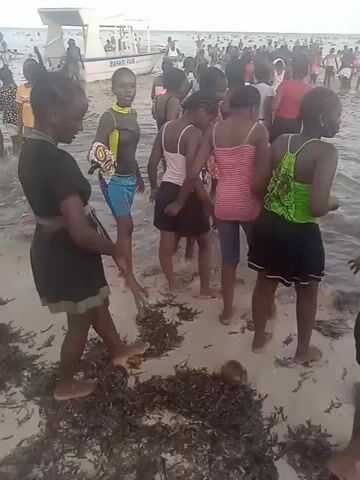 A group of children, mostly girls, wade in shallow water at a beach. Some are wearing swimwear and others are wearing casual clothing. The water is murky with seaweed and algae. In the background, there are boats and more people in the water. The children appear to be enjoying themselves and playing in the water.