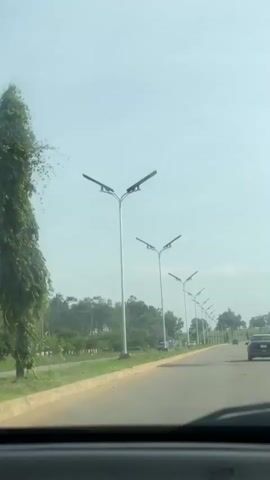 The video shows a view from inside a car driving down a paved road. On the left side of the road, there is a grassy median strip and a row of modern streetlights. The streetlights are tall and white, with two angled arms each supporting a rectangular light fixture. Beyond the median is a line of trees. On the right side of the road, there is another row of similar streetlights receding into the distance. A few cars are visible on the road ahead. The sky is overcast and pale blue. The perspective suggests the video was filmed by someone in the passenger seat or from the dashboard.