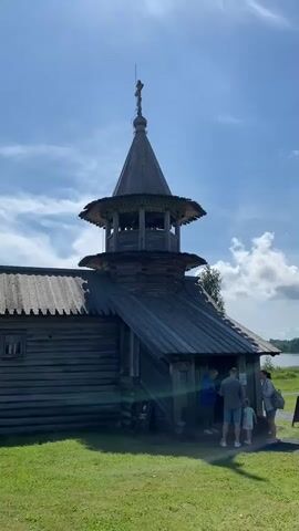 The video shows an old wooden church with a bell tower. There are several people standing in front of the church entrance, and a child is with them. The weather appears to be sunny with some clouds in the sky. The church is surrounded by a green grassy area. There is a body of water visible in the background. The video appears to be a short clip of tourists visiting a historical site.