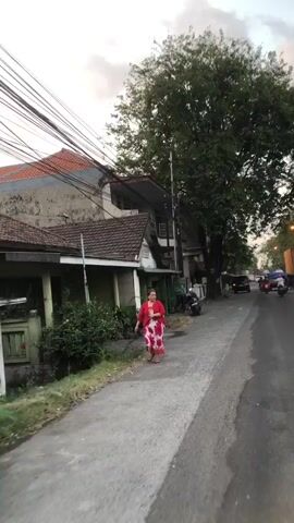 A woman in a red and white patterned dress walks along a paved sidewalk next to a street lined with trees and buildings. A motorcycle is parked on the side of the road. The sky is overcast.