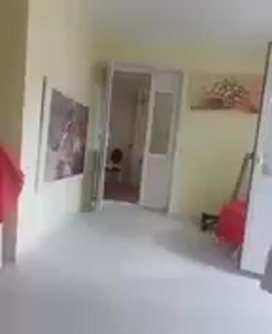 The video shows an interior view of a room. The walls are painted a light yellow color. On the left wall, a framed picture is hanging. Across the room, a doorway is open, revealing a small space beyond, possibly a bathroom, with a round object visible on the floor. Another framed picture hangs on the right wall. In the foreground, on the right side of the frame, a white cabinet or piece of furniture is partially visible, covered with a light-colored tablecloth. A red cloth is draped over an object leaning against the right wall, and another red object is visible in the background to the left of the open doorway. The floor is a light, neutral color. The overall impression is of a simple, sparsely furnished room.
