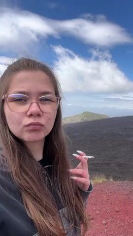 A young woman stands outdoors in front of a volcanic landscape under a blue and cloudy sky, taking a selfie video. She has long brown hair and wears eyeglasses and a gray sweatshirt. She holds a lit cigarette in her right hand with long white nails, which she brings to her mouth to smoke. In the background, there is a barren, dark volcanic landscape, a distant view of a valley, and a sky with scattered clouds. The ground near her feet is reddish in color.
