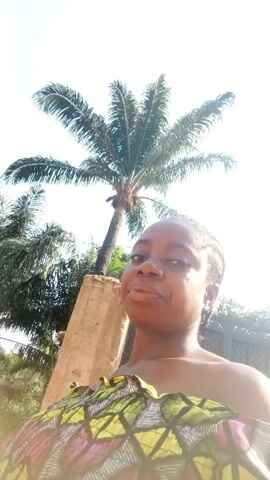 A person is standing outdoors in front of a palm tree. The sun is bright and the sky is white. The person is wearing a colorful patterned fabric. A cement block is to the person's left. A fence is behind the person. The person has short dark hair and is looking directly at the camera with a neutral expression. The camera is held low, angled up towards the person.