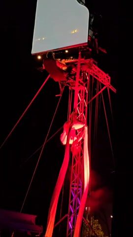 At night, an aerial performer hangs suspended from red fabric straps attached to a metal truss structure. The performer is in a bent-knee, spread-leg position, with arms extended, as they move. At the very top of the truss, a person is seated at a white grand piano which is illuminated from below by red stage lights. The piano is mounted on a platform atop the truss. Red cables crisscross the scene, connecting various points of the truss and possibly the piano. The background is completely black, indicating it is nighttime.
