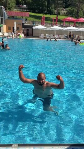 A bald man with a beard is in a swimming pool. He is flexing his arms and then splashes the water. He is wearing black swimming shorts. In the background, there are other people in the pool, lounge chairs, umbrellas, and a building. The water is bright blue.