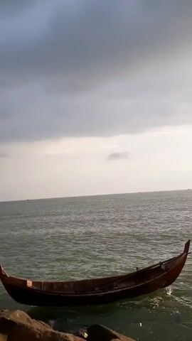 The video shows a man sitting on a large rock facing the sea and using a mobile phone. He is wearing a black t-shirt and light-colored shorts. To his right, a traditional boat with red trim sits at the edge of the water. The sky is overcast with dark, looming clouds. Further down the rocky shoreline, a few other people are visible, standing near the water's edge. The ocean is calm with slight ripples.