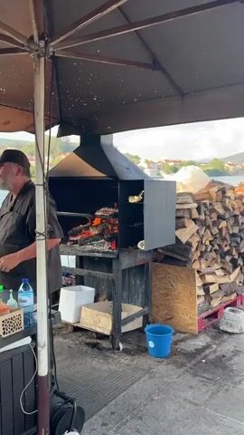 A man in a brown apron stands next to a large outdoor grill with a fire burning inside. To the right of the grill, there is a large pile of chopped firewood stacked on pallets. In the background, a body of water is visible, with buildings and trees on the opposite shore. The sky is cloudy. A man in a red shirt stands with his back to the camera looking out at the water.