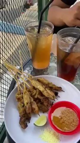 The video shows two people eating a meal outdoors. On the table, there are two plates of food: one with satay skewers, a wedge of lime, a small red bowl of peanut sauce, and a small pile of green powder; the other with rice, peanuts, and skewered meat topped with sauce. There are also two glasses of iced tea with straws. One person is visible in the frame, wearing a black t-shirt with a beach-themed design. They are using a spoon to pour sauce over the food on their plate. The background shows a green metal fence and a blurry view of the street outside.