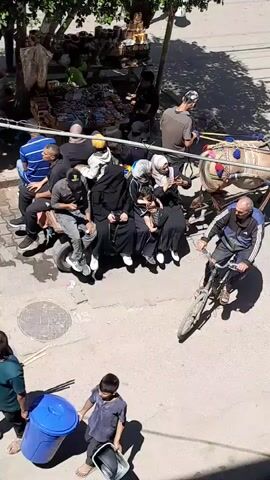 The video shows a busy street scene with a donkey-drawn cart carrying several people. The cart appears to be made from a repurposed vehicle chassis, with a single wheel on each side and a wooden plank for seating. The people on the cart are of various ages and genders, some wearing traditional Islamic clothing. They are seated closely together, with some standing behind those seated. A man on a bicycle is seen riding alongside the cart, and another person is walking in the background. There are also market stalls visible on the side of the street, suggesting a commercial area. The overall impression is of a community using traditional and makeshift transportation in a lively urban environment.