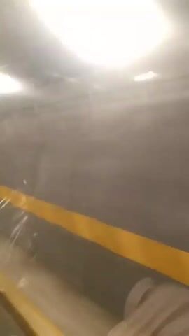 From a low angle inside what seems to be a moving railcar, the camera records the blurred view of an interior wall and a yellow safety stripe, indicating the edge of the floor. The wall is a neutral, gray color. Light from above is casting a bright reflection off of the wall. Occasional movement and shadows indicate the possibility of people and objects just outside the frame, but it is mostly a blurred and obscured view of the railcar's interior.