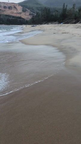 The video shows a sandy beach, with a gentle wave washing over the sand in the foreground. In the distance, there are mountains covered in trees, along with what appears to be a coastal road or highway. A small boat sits on the beach. A person can be seen walking on the beach further down the shoreline. The sky is overcast, providing a muted light across the scene. The video is stable, providing a clear view of the beach.