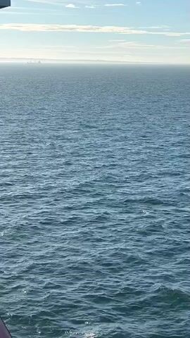 The video shows a vast expanse of blue ocean under a partly cloudy sky. The water is choppy with visible waves. In the far distance, a hazy line of land or a cityscape is visible on the horizon. The bottom left corner of the frame shows a dark, possibly red, railing, suggesting the viewer is on a ship or a balcony overlooking the water. The lighting indicates it is daytime, likely morning or afternoon, with the sun creating a glare on the water. No people are visible in the frame.