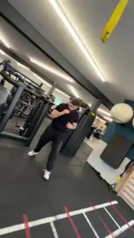 A man in a black shirt and pants with white sneakers is shadow boxing in a gym. The gym is well-lit with various exercise machines and equipment visible. The man has a beard and tattoos on his arm. He is facing the camera. The camera angle moves around him while he is boxing.