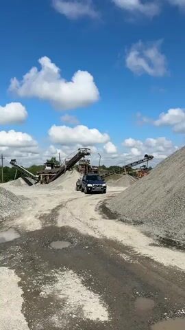 A dark blue SUV with a small trailer is driving on a dirt road. The road is surrounded by piles of gravel. In the background, there is industrial machinery, possibly a rock crusher, with conveyor belts. The sky is blue with white clouds.