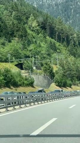 The video shows a highway with traffic moving slowly or stopped. The road is paved and has white lane markings. A metal guardrail separates the lanes of traffic from the side of the road. On the side of the road, there is a steep, tree-covered mountain slope. Some trees are tall and evergreen, while others are shorter and deciduous. There are also power lines and poles visible on the mountainside. A few cars are visible on the highway, including a black SUV with a roof rack, a white van, and a black Mercedes-Benz sedan. The sky is not visible in the frame. The overall impression is a scenic mountain highway experiencing traffic congestion.