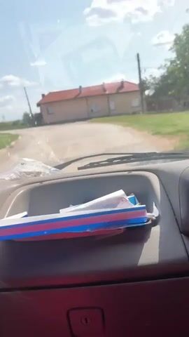 The video shows a view from inside a vehicle, looking through the windshield. The dashboard of the vehicle is visible in the foreground, with a few items on it, including a blue and red striped folder or binder and some papers. A can, possibly a beverage can, is in a cup holder to the left. Outside the vehicle, the road curves ahead, with a light-colored building with a red-tiled roof on the right side of the road. Further down the road, another building with a red-tiled roof is visible. The sky is blue with some clouds. The scene appears to be a rural or suburban setting. The video is short, with a duration of about 4 seconds, and the camera is static, not panning or zooming.