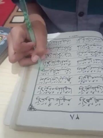 A person's hand is holding a green pen and writing in a book. The book is open to a page with Arabic text. The person is wearing a grey and green striped shirt. The background is blurred but appears to be a wooden surface and possibly other books. The lighting suggests it is daytime, indoors.