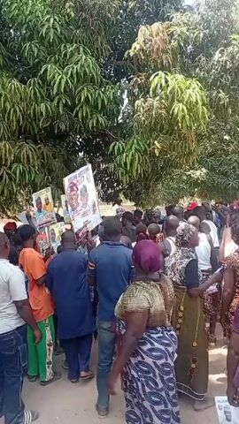 A large crowd of people, mostly African, are gathered in an outdoor area with trees overhead. Many of them are holding signs, presumably for a political campaign, with portraits of men in suits. The signs read "Value Abaji Area 2027". Some people are wearing traditional African clothing. The crowd is moving around, possibly marching or protesting. The sounds of drumming and music are audible in the background.