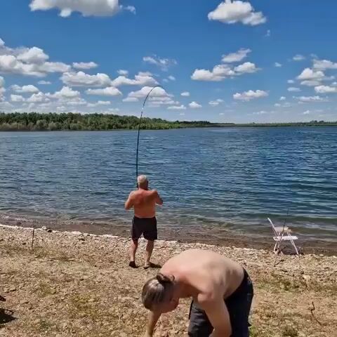 Two shirtless men are by a lake. One man, closer to the camera, is facing away and appears to be reaching down or adjusting something. The other man is further away, standing at the water's edge and holding a fishing rod, which is bent, indicating a fish may be on the line. A white fishing rod holder is visible on the right side of the frame. The environment is a shoreline with gravel and some dry vegetation, with a large body of water in the background and a tree line on the opposite shore. The sky is blue with scattered white clouds.