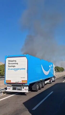 An Amazon Prime branded semi-trailer truck is visible on a highway under a clear blue sky. Dense plumes of dark smoke are rising in the distance, obscuring the horizon and suggesting a fire. The truck appears to be stationary, as do other vehicles on the highway, indicating a potential traffic obstruction or incident related to the smoke. The rear of the trailer is white with text that includes "Shipping. Streaming. Savings. It's on amazon prime." The side of the trailer is bright blue with the Amazon smile logo. The truck is on the right side of the frame.