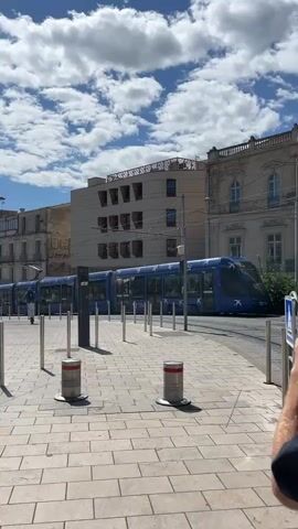 A blue tram glides to a stop on its tracks in a city environment. The camera is situated on a paved walkway, bordered by a series of metal bollards, some with red and silver tops. To the left, a person in dark clothing walks away from the tram, carrying a white shopping bag. In the background, there's a blend of building styles, from older structures with intricate facades to more modern buildings with geometric designs. Above, a partly cloudy sky suggests a bright day. The walkway is constructed from square paving stones, and the scene is well-lit by natural sunlight. In the background, metal security gates create a frame around some greenery.