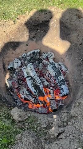 A close-up, overhead view of a fire pit dug into the ground. The pit contains burning logs and glowing embers, with flames visible. The edges of the pit are made of dirt. Shadows of two people are visible at the top of the frame. The ground around the pit is covered in grass.