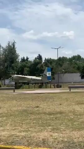 The video shows a parked, pale green or light blue twin-propeller aircraft with "ARMADA" written on its side, which the camera is driving past. The plane is on a concrete display stand in a grassy field, with trees and bushes in the background. There is also a road with a white sedan driving on it. The sky is mostly blue with some white clouds. The video is taken from inside a car, looking out the window.