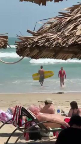 The video shows a beach scene. In the foreground, several people are relaxing on beach chairs under thatched umbrellas. In the background, a man is walking into the ocean carrying a yellow bodyboard, followed by a woman. The ocean has small waves. The sky is bright.