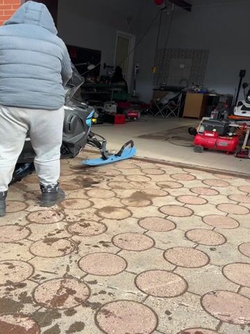A person in a grey puffy jacket and grey sweatpants is pulling a blue and black snowmobile out of a garage. The garage is cluttered with tools, a motorcycle, and other equipment. The ground outside the garage is made of patterned pavers. The person is walking backwards while pulling the snowmobile.