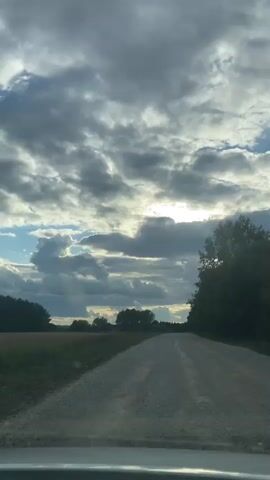 The video shows a view from inside a vehicle traveling on a gravel road through a rural landscape. The sky is filled with dramatic, gray, and white clouds, with patches of blue sky visible and the sun shining through in the distance. The road stretches ahead, bordered by fields and trees on either side. The vehicle appears to be moving forward, indicated by the slight blur of the surroundings and the perspective of the road. The overall impression is one of natural beauty and the journey through a serene, possibly overcast, rural environment.