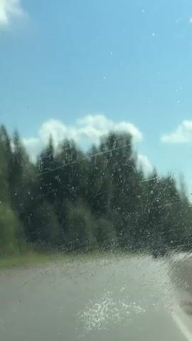 The video is a first-person perspective recording from inside a vehicle. The windshield is visible in the foreground with water droplets and streaks on it, suggesting it might be raining or has recently rained. A windshield wiper is also present. The video shows a road curving to the right in the middle ground, with a dark-colored car driving ahead on the road. Lush green trees line the sides of the road and extend into the background. The sky above is blue with scattered white clouds. The overall impression is that of a scenic drive on a clear day with some residual moisture on the windshield.