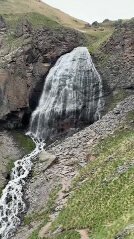 The media file shows a wide, multi-tiered waterfall cascading down a rocky mountainside in a natural, outdoor setting. The waterfall is the central focus, with water flowing from a broad crest and fanning out as it descends. The surrounding landscape is characterized by rugged, dark rock formations and slopes covered in patches of green grass, indicating a mountainous region. The sky appears overcast, suggesting a cloudy or possibly foggy day, which casts a diffused light on the scene. The overall impression is one of natural grandeur and remoteness, typical of a mountainous terrain.

The primary subjects are the waterfall and the natural landscape it inhabits. There are no visible people or man-made objects, except for a small yellow marker on a hiking path visible in the lower right portion of the frame, suggesting the location might be a hiking destination. The activity is the continuous flow of water over the rocks, a natural phenomenon.

Notable details include the sheer volume of water in the waterfall, the textured rock faces, and the contrasting green vegetation clinging to the slopes. The weather appears to be cool and overcast, adding to the dramatic and somewhat somber atmosphere of the rugged environment. The location cues, such as the type of vegetation and the rocky, mountainous terrain, are consistent with a location like Terskol, Russia, known for its mountainous landscapes and natural features. There is no visible text in the media file, other than the yellow marker.