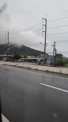 The video shows a wet highway with a concrete barrier on the right. There are power lines and poles on the left, with a misty mountain in the background. Several trucks are driving on the highway, including one with a red shipping container that says "tex" and another with a brown shipping container that says "HMM". The road is wet and appears to be raining, as indicated by the spray from the truck tires. The camera appears to be mounted in a vehicle, possibly a car or bus, looking out the window.