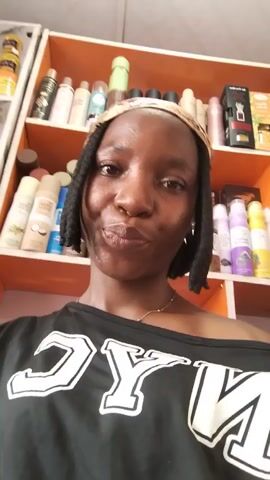 The video shows a young Black woman with braided hair, wearing a black t-shirt with white "NYC" text. She is in a room with shelves filled with various products, possibly cosmetics or toiletries. The shelves are orange and white. The woman is looking at the camera and making faces. The lighting suggests it is daytime. The camera is held at eye level, and the woman is close to the lens.